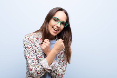 Genç, güzel bir kadın sevinçle gülümsüyor ve kollarını kavuşturup, yumruklarını sıkıp, mavi duvara karşı mutlu ve pozitif şeyler söylüyor.