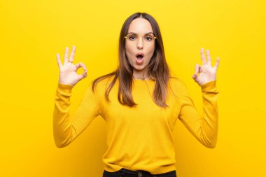 Genç, güzel bir kadın şok olmuş, şaşırmış ve şaşırmış hissediyor. Onaylıyor. İki eliyle turuncu duvara işaret veriyor.