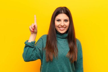 Genç, güzel bir kadın mutlu ve mutlu bir şekilde gülümsüyor. Bir eliyle sarı duvara uzayı yansıtıyor.