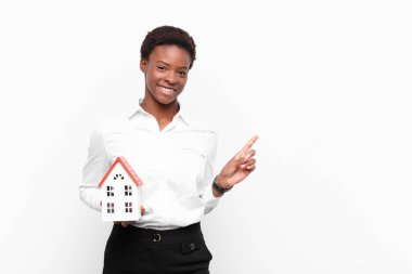 Genç, güzel siyah kadın neşeyle gülümsüyor, mutlu hissediyor ve yan ve yukarı doğru işaret ediyor, bir ev modeliyle fotokopi odasında nesneleri gösteriyor.