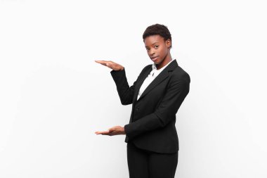 Genç, güzel, siyah bir kadın, iki eli yanda olan bir objeyi tutuyor. Fotokopi alanı, beyaz duvara karşı bir objeyi gösteriyor, sunuyor ya da reklamını yapıyor.