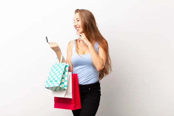 若いブロンドの女性は幸せな自信のある表情で笑顔で顎を手にショッピングバッグを持っている側に疑問と見て — ストック写真