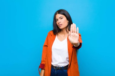 Genç, güzel, Latin bir kadın ciddi, sert, hoşnutsuz ve kızgın görünüyor. Avuç içini açıp düz duvara karşı jest yapıyor.