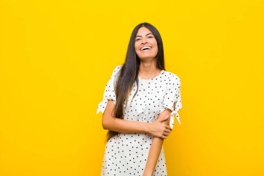 Genç ve güzel Latin kadın utangaç ve neşeli bir şekilde gülüyor. Düz duvara karşı dostane, olumlu ama güvensiz bir tavır takınıyor.