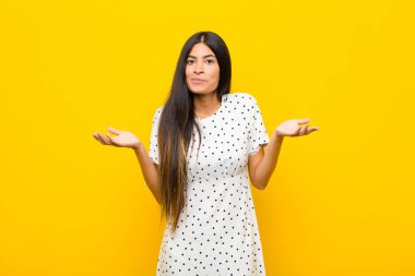 Genç, güzel, Latin bir kadın şaşkın ve şaşkın, şüpheci, farklı seçenekleri tartıyor ya da düz duvara karşı komik bir ifadeyle seçiyor.