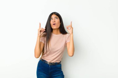 Genç, güzel, Latin bir kadın şaşırmış, şaşırmış ve ağzı açık görünüyor, iki eliyle yukarıya bakıyor düz duvara uzayı kopyalamak için.