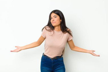 young pretty latin woman feeling clueless and confused, having no idea, absolutely puzzled with a dumb or foolish look against flat wall