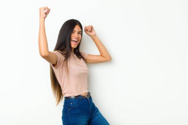 Genç, güzel, Latin bir kadın muzaffer bir şekilde bağırıyor, heyecanlı, mutlu ve şaşırmış görünüyor, düz duvara karşı kutlama yapıyor.