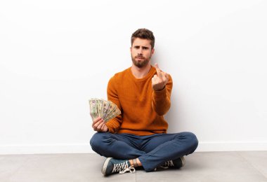 Genç ve yakışıklı bir adam kızgın, sinirli, asi ve agresif, orta parmağını sallıyor, dolar banknotlarına karşı koyuyor.