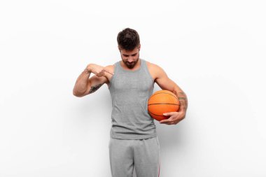 Genç yakışıklı adam neşeyle gülümsüyor, yere bakıyor ve elinde bir basketbol topu tutarak göğsünü işaret ediyor..
