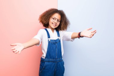 Afro-Amerikan küçük kızı neşeyle gülümsüyor sıcak, dostça, sevgi dolu bir kucaklama yapıyor, mutlu ve sevimli hissediyor düz duvara karşı.