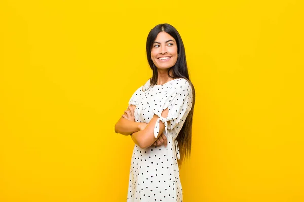 young pretty latin woman smiling gleefully, feeling happy, satisfied and relaxed, with crossed arms and looking to the side against flat wall