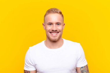 Genç sarışın yakışıklı adam mutlu ve şapşal görünüyor. Geniş, eğlenceli, kaçık bir gülüşü ve sarı duvara bakan gözleri var.