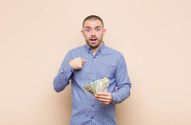 Genç ve yakışıklı adam şok olmuş ve şaşırmış görünüyor, ağzı sonuna kadar açık, dolar banknotlarıyla kendini işaret ediyor.