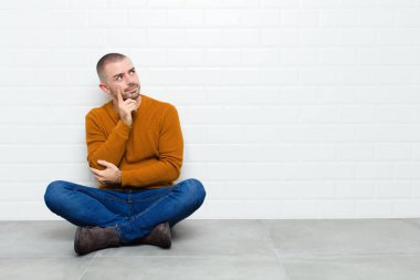 Genç ve yakışıklı bir adam konsantre bir bakışla, şüpheli bir ifadeyle, yukarı ve yana bakıyor ve yerde oturuyor