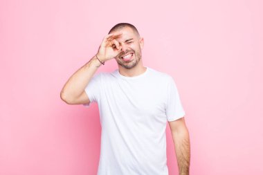 Genç yakışıklı adam mutlu bir şekilde gülümsüyor, şaka yapıyor ve gözetleme deliğinden bakıyor, düz duvara karşı sırları gözetliyor.