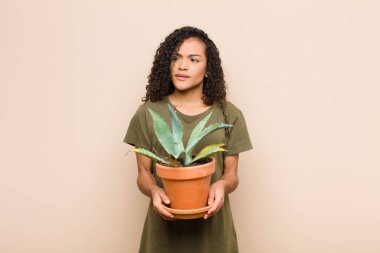 Genç siyahi kadın şaşkın ve kafası karışmış hissediyor. Kafasını kaşıyor ve elinde kaktüsle yan tarafa bakıyor.