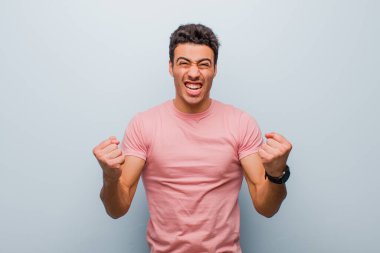 Genç Arap adam sinirli, sinirli, sinirli bakışlar ve yumruklarla bağırıyor, gri duvara karşı öfkeli hissediyor.