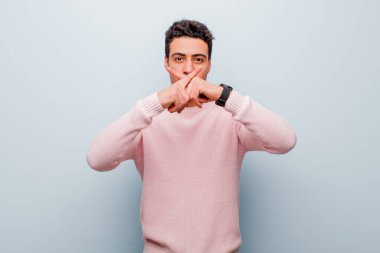 Genç Arap adam ciddi görünüyor ve iki parmağının önde olması hoşnutsuzca reddedilerek gri duvara karşı sessizlik istiyor.