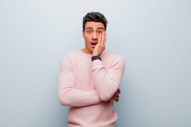 Genç Arap adam şok ve inançsızlıkla ağzı açık, elleri yanak ve kolları çapraz, sersemlemiş ve gri duvara karşı şaşkın hissediyordu.