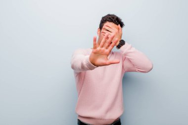 Genç Arap adam elini yüzünü örtüyor ve diğer elini kamerayı durdurmak için öne koyuyor. Gri duvara karşı fotoğraf ya da resim çekmeyi reddediyor.