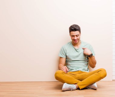 Genç Arap adam neşeyle gülümsüyor, yere bakıyor ve göğsünü işaret ediyor.