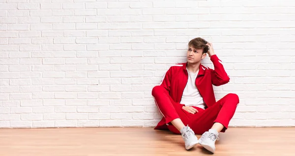 young blonde man feeling puzzled and confused, scratching head and looking to the side sitting on the floor