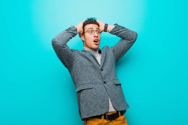 Korkunç bir hata yüzünden dehşete düşmüş ve şok olmuş görünen ağzı açık genç bir Arap adam mavi duvara karşı el kaldırıyor.