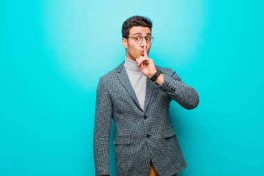 Genç Arap bir adam sessizlik ve sessizlik istiyor, parmaklarıyla ağzının önünde işaret ediyor, ya da mavi duvara karşı bir sır saklıyor.