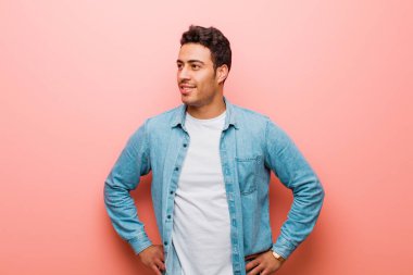 Genç Arap adam mutlu, neşeli ve kendinden emin görünüyor, gururla gülümsüyor ve iki elini de pembe duvara yaslamış kalçalarını kolluyor.