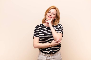 Orta yaşlı bir kadın mutlu, kendinden emin bir ifadeyle gülümsüyor, elleri çenesinde, merak ediyor ve yan tarafa bakıyor.