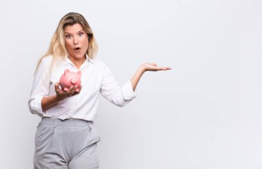 Genç ve güzel Latin kadın şaşırmış ve şoka uğramış görünüyor, çenesi düşmüş, eli açık bir objeyi elinde tutarken beyaz duvara karşı bir kumbara