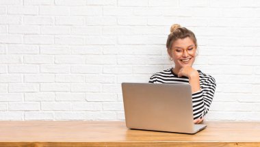 Genç ve güzel Latin bir kadın mutlu, kendinden emin bir ifadeyle gülümsüyor, çenesinde elleri, merak ediyor ve yan tarafa bakıyor dizüstü bilgisayarıyla oturuyor.