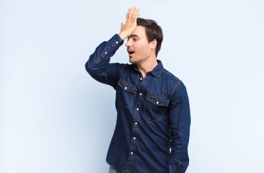 young handsome man raising palm to forehead thinking oops, after making a stupid mistake or remembering, feeling dumb against blue wall