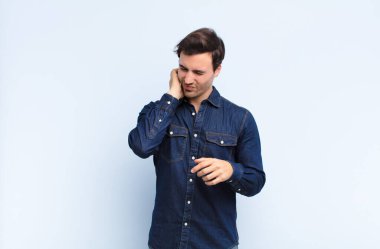 Genç yakışıklı bir adam stresli, hayal kırıklığına uğramış ve yorgun hissediyor. Endişeli, sorunlu bir bakışla mavi duvara yaslanmış boynu okşuyor.