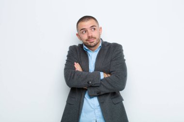 Genç işadamı omuz silkiyor, kafası karışık ve kararsız, kolları çapraz ve şaşkın bir şekilde düz duvara bakıyor.