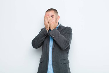 Genç işadamı elleriyle yüzünü kapatıyor, parmaklarının arasından şaşkın bir ifadeyle bakıyor ve düz duvara bakıyor.