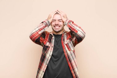 Genç ve yakışıklı bir adam stresli ve endişeli hissediyor, depresyonda ve hayal kırıklığına uğramış, baş ağrısı ile iki elini kaldırarak düz duvara karşı