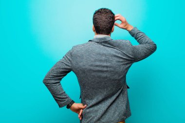 Genç Arap bir adam bilgisiz ve kafası karışık hissediyor, bir çözüm düşünüyor, eli kalçasında, diğeri kafasında, mavi duvara karşı dikiz manzaralı.