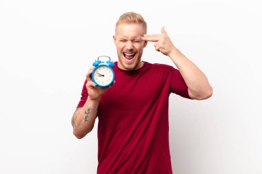 Genç sarışın adam mutsuz ve stresli görünüyor, intihar jesti elle silah işareti yapıyor, kafasını beyaz duvara doğrultuyor, elinde çalar saat var.