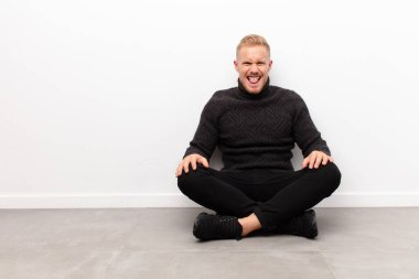 Genç sarışın adam tiksinmiş ve rahatsız olmuş hissediyor, dilini dışarı çıkarıyor, iğrenç bir şeyden hoşlanmıyor ve çimento zeminde oturuyor.