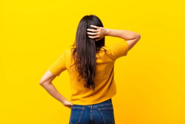 Genç Latin güzel kadın düşünüyor ya da şüphe ediyor, kafasını kaşıyor, kafası karışık ve kafası karışık, düz duvara karşı arka ya da arka pencereden bakıyor.