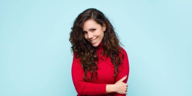 Genç ve güzel bir kadın utangaç ve neşeli bir şekilde gülüyor, dost canlısı ve pozitif ama güvensiz bir tavırla mavi duvara karşı.