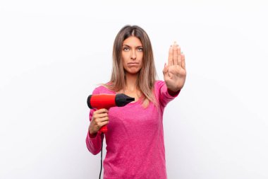 Genç ve güzel bir kadın ciddi, sert, hoşnutsuz ve kızgın görünüyor. Avuç içini açıp saç kurutma makinesiyle jest yapıyor..