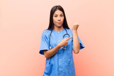 Genç Latin hemşire sabırsız ve kızgın görünüyor, saati gösteriyor, dakiklik istiyor, pembe duvara karşı zamanında olmak istiyor.