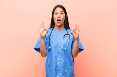 Genç Latin hemşire şok olmuş, şaşırmış ve şaşırmış hissediyor. İki elini de pembe duvara yaslayarak onay veriyor.