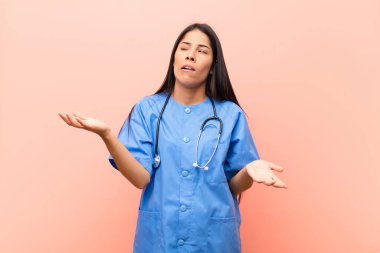 young latin nurse shrugging with a dumb, crazy, confused, puzzled expression, feeling annoyed and clueless against pink wall