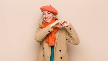 Yaşlı ya da orta yaşlı güzel bir kadın gülümsüyor ve mutlu, sevimli, romantik ve aşık, iki eliyle kalp şekli veriyor.