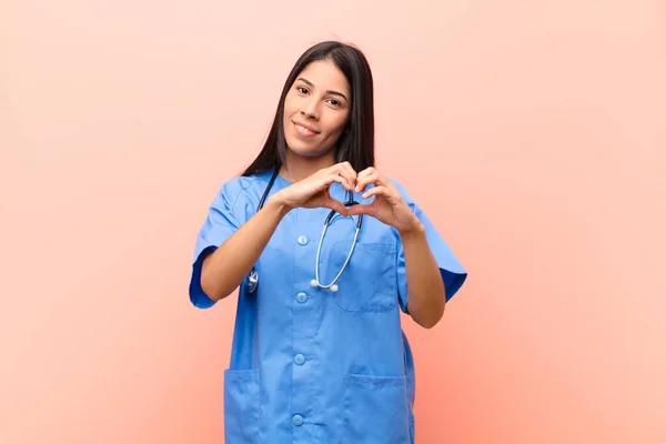 young latin nurse smiling and feeling happy, cute, romantic and in love, making heart shape with both hands against pink wall
