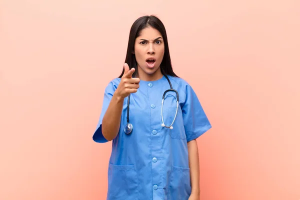 young latin nurse pointing at camera with an angry aggressive expression looking like a furious, crazy boss against pink wall